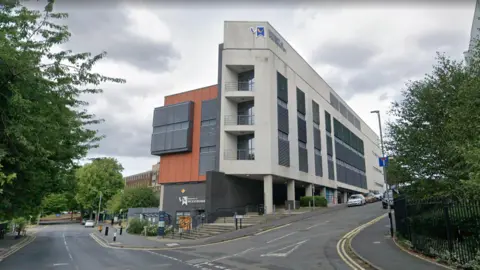 Google A concrete and grey-clad university building sits on a street corner, with an orange panel section on the upper left side. The building has several storeys with dark louvred windows and the University of Wolverhampton logo and branding visible on the facade. Trees line the road to the left, with parked cars visible on the street to the right.
