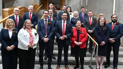 Group of around 15 people wearing business attire and red floral lapel pins pose for a photograph on a staircase inside a modern parliamentary-style building. The group is arranged across several steps, with handrails, concrete walls and wooden architectural features visible in the background.