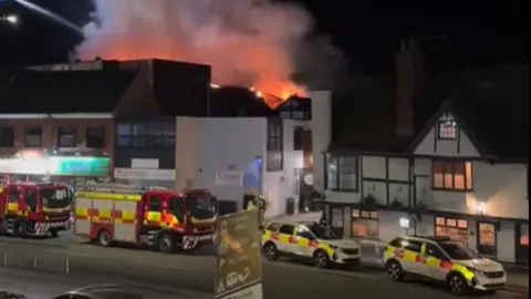 Fire service vehicles parked by the side of a road. A fire can be seen in the background with flames and smoke.