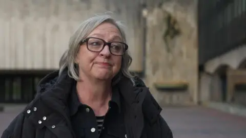 Paula Maines has shoulder length grey hair and black-rimmed glasses. She is wearing a black coat and is pictured outside Newcastle Civic Centre. 
