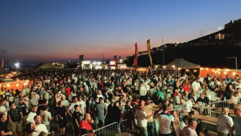 Hundreds of football fans fill a seafront fan park in Brighton at dusk, with food stalls, tents and large screens visible beneath a clear evening sky.
