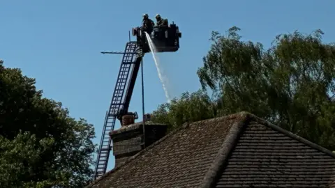 Firefighters operate an aerial ladder platform above a building roof, directing a jet of water downwards. The extended ladder rises above nearby trees, illustrating the specialist equipment used to tackle fires from height.