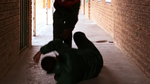 Getty Images A boy on the floor in a corridor raising his arm to defend himself as another boy who is poised to hit him is standing above him