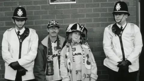 A black and white image of a male and female football fan wearing hats, badges, scarfs and football shirts. He has his arm around her. The are standing between two West Midlands Police officers who have helmets, and uniform on. They are both wearing black gloves and have their hands crossed in front of them. 