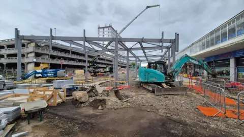 Stuart Woodward/BBC Harlow bus station under reconstruction, photographed on 11 May 2026. A frame of steel girders is in place, with a crane in the background, diggers in the foreground and other construction materials as well