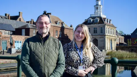King's Lynn and West Norfolk Borough Council Simon Ring is wearing a dark green body-warmer over a lighter green top. He is smiling and standing next to a woman who is wearing a leopard print, long-sleeve top. Behind them is the Custom House building.
