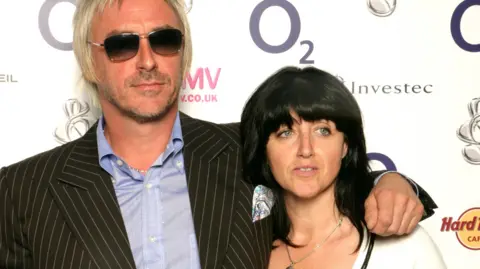 A man with platinum blond hair with sunglasses and wearing a black suit and light blue shirt, standing next to a woman with black hair and a white shirt.