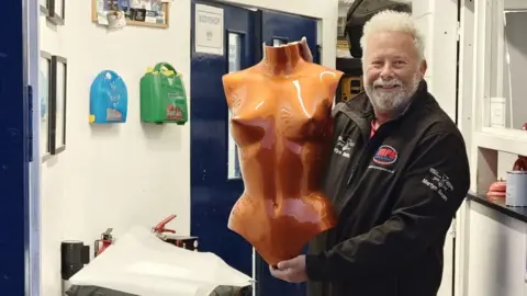 MPS Body & Paint Martyn Smith holds up a metallic orange breastplate. He is smiling and stood in a corridor, in front of a blue door labelled bodyshop.