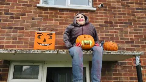 A fake plastic figure wearing jeans and a hoodie and a plastic mask holding a pumpkin. He is perched on a porch outside a home.