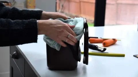 West Northamptonshire Council A woman's hands, on a black food caddy, with a peeler on a white counter with two carrots by some knives. The woman has painted nails, two rings on her fingers and is wearing a dark top 