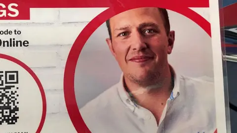 Stephen Huntley/BBC A poster detailing information about heart screenings with the image of a man who died. He has short dark hair and wears a white shirt in the image. 