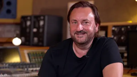 Nick Brine now. He has chin-length dark hair, slicked back, a stubbly beard and is wearing a black t-shirt with the recording studio desks blurred in the background.