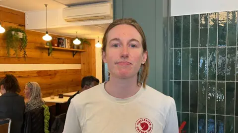 Holly Nichols/BBC Roisin McNeill standing inside a cafe with a green tiled wall behind her on the  right and three people sitting at tables behind her on the left. She has light brown hair pulled back from her forehead and neck and is wearing a white round-necked T-shirt. 