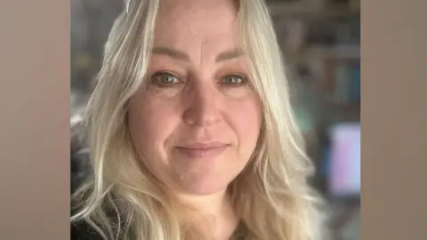 Michelle Bullivant Michelle Bullivant who has long blonde hair over her shoulders. She is looking at the camera and a dark top can be glimpsed.