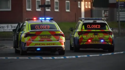 Two police cars marked yellow, orange and blue with flashing blue and red lights parked on a road at a junction facing away from the camera. There is a blue and white police cordon between the cars and the camera.