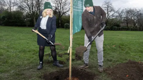 Duncan Lomax, White Rose Forest Tracy Brabin and David Skaith wear green wooly hats to plant an Oak Tree sapling. They hold spades.