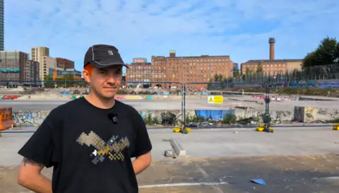 BBC A young man wearing a black t-shirt and cap stands with his arms behind his back. He's standing in front of the demolished skatepark, which is cordoned off by a fence. The park is strewn with rubble and litter.