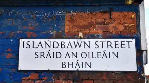 A white bilingual street sign in Irish and English. It is on a red brick wall that is partially painted blue. Some barbed wire can be seen on the wall above the sign. 
