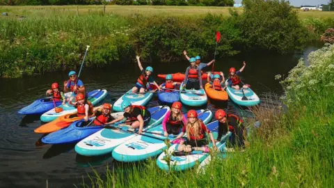 Mendip Adventure A group of young people smile and wave to the camera as they sit in kayaks on a river.