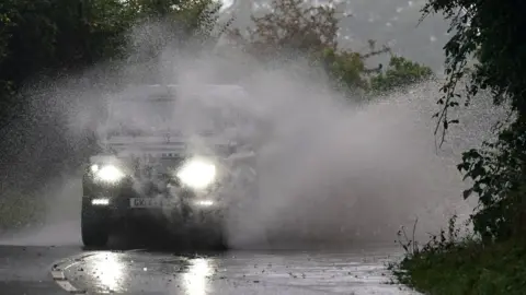 PA Media A jeep-style vehicle causes a huge wall of spray to fly up as it drives on a flooded road near Gloucester.