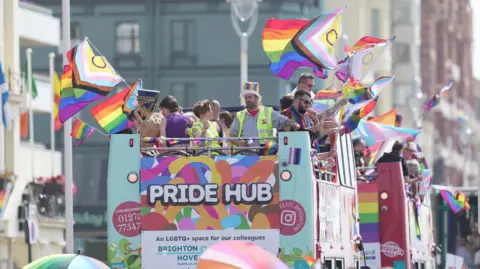 Eddie Mitchell Pride Hub buses making their way along Kings Road. People are stood on the top deck of the open top buses waving rainbow flags