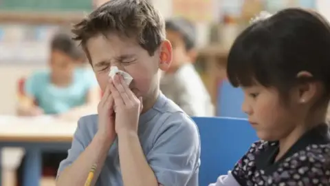Stock photo of boy in class blowing his nose. He has short brown hair and a grey t-shirt. He is sat next to  girl with dark hair and a black and white patterned top. 