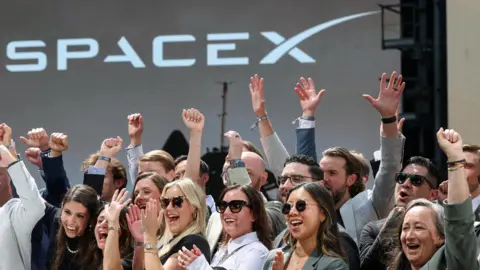 Group of people in the foreground raising their arms and clapping in front of a large stage backdrop displaying the SpaceX logo.