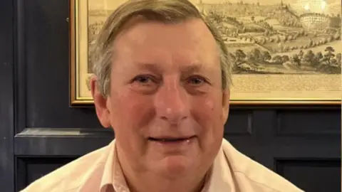 A man with grey hair wearing a white shirt smiles at the camera. He is sitting in a room with black panelling and an old-fashioned drawing on the wall.