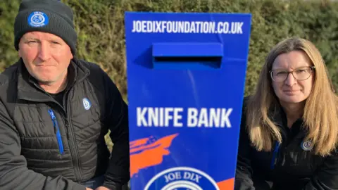 Phil and Emma Dix crouch either side of a blue rectangular bin with a letter box-style opening. Signage on the bin reads joedixfoundation.co.uk and Knife Bank. Phil Dix is on the left of the image and wearing a black jacket and black hat. Emma Dix, to the right, is wearing the same black jacket. She has long fairish hair and glasses. Both are looking at the camera. 