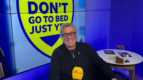 A man with grey and thick black-rimmed spectacles sits in a blue-lit recording studio. A blue and yellow badge with the words 'Don't go to bed just yet' is on the wall behind him. A BBC Radio Leeds mic is in front of him.