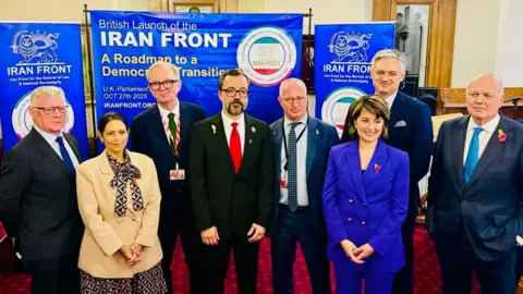 Mattie Heaven Mattie Heaven is wearing a blue trouser suit and her husband, Vahid Behesti is standing in the middle of the picture, wearing a dark suit and bright red tie. Both are surrounded by UK parliamentarians at the Palace of Westminster, including the former home Secretary Priti Patel and former Conservative leader Iain Duncan-Smith.