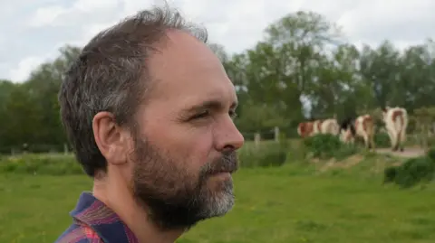 John Fairhall/BBC Jonny Crickmore stands in a grass field and looks away from the camera. He has short dark hair and a thick dark beard. He wears a red and blue chequered shirt. His herd of cattle can be seen in the distance.