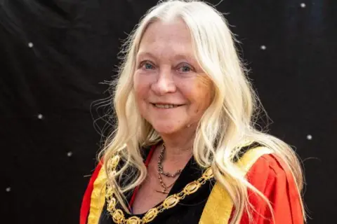 Nantwich Town Council A woman with long light blonde hair, wearing a read mayoral robe and gold chain around her neck, stands and smiles in front of a black sheet with white spots.