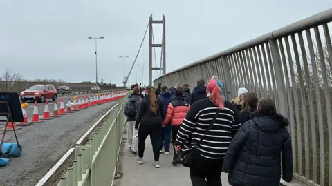 Jake Zuckerman / BBC Sponsored walk participants with their backs to the camera walk across the Humber Bridge