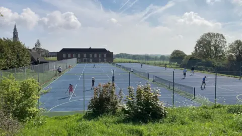 Six tennis courts in a line, with the Royal Hospital School's clock tower and a boarding house at the far end.