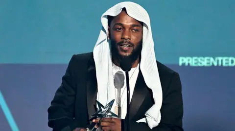 Kendrick Lamar on stage with a white hood and black jacket, holding his award