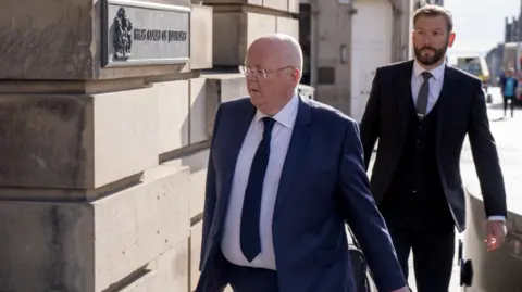 PA Media Two people in dark business suits walk along a sunlit pavement beside a stone building. The person in front is Peter Murrell and is wearing glasses and carrying a bag, passes a wall plaque reading “High Court of Justiciary.” A second suited person follows a few steps behind. The background shows a bright street scene with buildings and pedestrians out of focus.