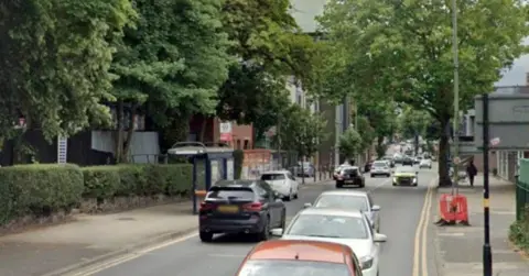 Traffic can be seen going in both directions on Kings Heath high street. There are also trees lining the street, with buildings behind them. A pedestrian can be seen walking in the distance.
