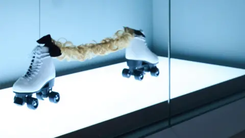 Simon Jacobs/St Albans Museum An art exhibit behind glass, which is lit from below. A pair of white roller skates with black wheels sit facing away from each other, with about 0.5 metres gap between them. They are joined by a curly blonde hair extension and two black bow ties.