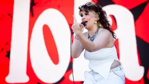 Getty Images Lola Young performing on stage with a finger raised, in front of a red and white backdrop saying "Lola"