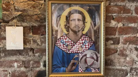 A framed illustration of a footballer styled like a religious icon hangs on a brick wall, with a halo around the head and the player wearing a red and white Croatia shirt while holding a football.