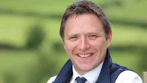 LMC Colin smiling. He has brown hair and is wearing a white shirt, blue tie and blue bodywarmer.