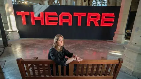 Richard Walker/ImageNorth A woman with shoulder length mousy brown hair with a serious expression looking upwards while sitting on a bench. In front of her are the words 'THEATRE' in large red letters on a black board.