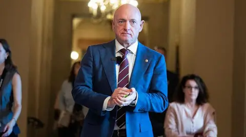 Senator Mark Kelly, wearing a suit, walking forward in a government building hallway with his hands clasped in front of him