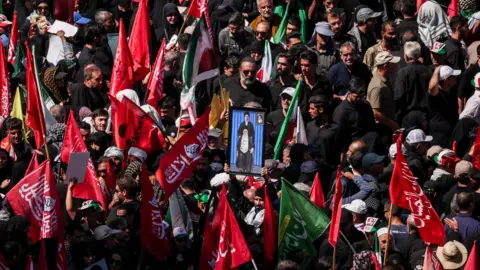 Reuters Mourners gather for the burial of Iranian Supreme Leader Ali Khamenei in Mashhad. Many are carrying red flags with Arabic script and in the centre a man holds a picture of a sitting Khamenei, with his son Mojtaba standing above him.