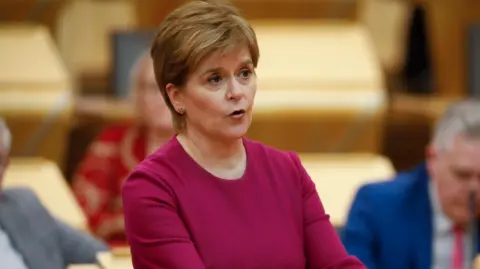 PA Media Nicola Sturgeon, who is wearing a maroon dress with a round neck, addresses MSPs in the Scottish parliament debating chamber. She has brown hair, with blond highlights.