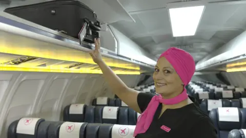 Chloe Jacquet Chloë Jacquet is dressed as an airline cabin crew, with a black dress, pink scarf and pink turban. She stands in the aisle of an empty plane and smiles at the camera as she touches a suitcase in an overhead cabin.