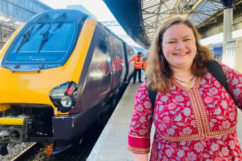 BBC Woman - Aimee Smith - in bright maroon dress, smiling at camera, on rail platform, with train in the background.