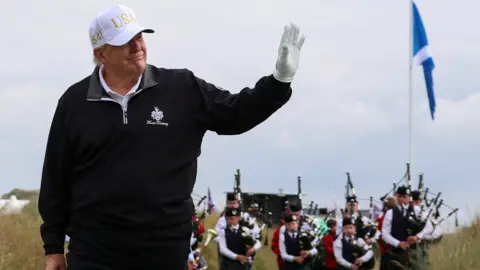 Reuters Donald Trump, wearing a black fleece, white USA cap and a white golfing glove, waves while walking on his golf course. Behind him is a saltire on a flag pole and a pipe band.