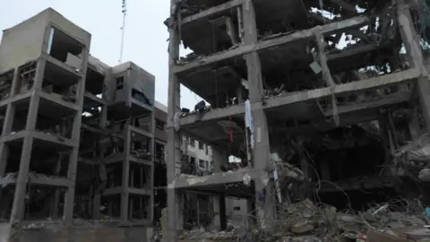 Two destroyed buildings, four to five stories high, with the windows and walls blown out. Some concrete pillars and floors remain. There are a few bits of fabric hanging off the structure and some twisted metal on the ground.